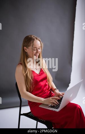 A happy business woman blogger smile in a red dress is working, typing on a laptop apple and sitting on a chair. portrait blonde assistant of hands with computer MacBook . Vertical Stock Photo