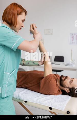 https://l450v.alamy.com/450v/2pmhbw7/redhead-physiotherapist-stretching-painful-arm-of-bearded-man-lying-on-massage-table-in-rehabilitation-centerstock-image-2pmhbw7.jpg