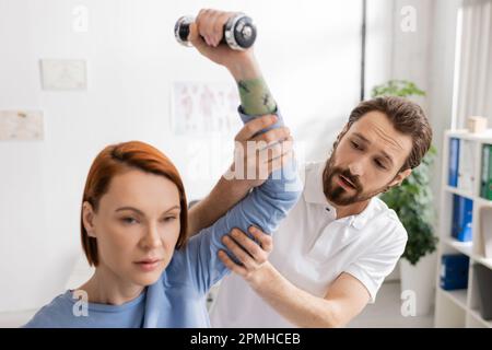 bearded physiotherapist supporting arm of redhead woman exercising with dumbbell in rehabilitation center,stock image Stock Photo
