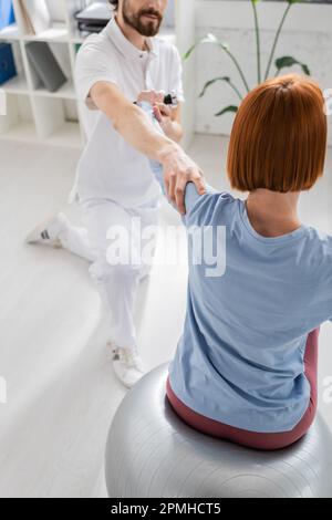 rehabilitologist supporting arm of redhead woman sitting on fitness ball and exercising with dumbbell in consulting room,stock image Stock Photo