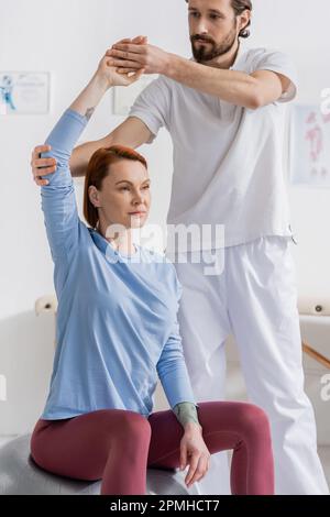physiotherapist supporting arm of redhead woman training on fitness ball in recovery clinic,stock image Stock Photo