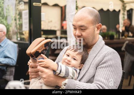 Flexible male infant looking at camera sitting on his dad's legs, holding his own legs, playing with father in beer garden at pub in Scotland. Joyful. Stock Photo