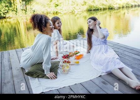 Happy female friends on picnic on the bank of a river. Blanket with pillows and food on a wooden bridge. Bottle of strawberry and lemon smoothie, soda Stock Photo