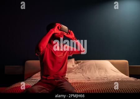 African man wearing VR glasses fully immersed in virtual world, sitting on bed under red neon light, Stock Photo