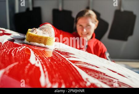 Young woman cleaning front hood of her car, closeup detail on yellow sponge held by hand in plastic glove Stock Photo