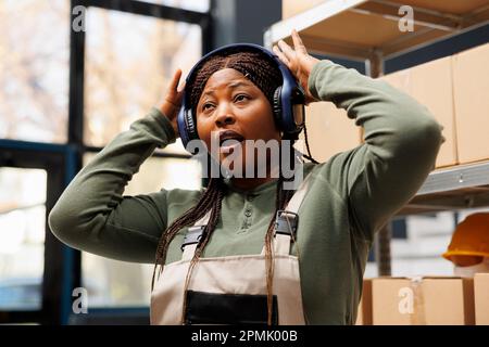 African american worker enjoying listening music in warehouse, working at customer packages before start shipping process. Stockroom employee wearing headset, dancing during work break Stock Photo