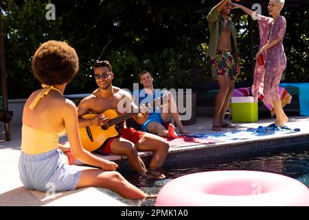 Happy diverse group of friends having pool party, playing guitar and dancing in garden Stock Photo