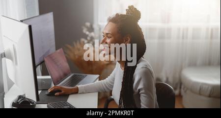 Smiled cheerful female web expert sitting at desk in living room coding on computer Stock Photo