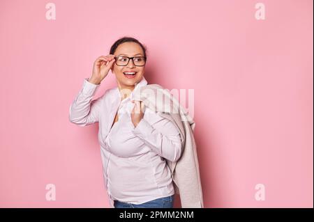 Pregnant cheerful ethnic woman smiling looking aside, putting trendy glasses on isolated pink background. Copy ad space Stock Photo