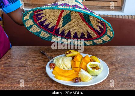 The waiter lifts a straw cover for food over a tropical fruits on a breakfast. Fresh banana, passion fruit, pineapple, mango, papaya, orange, dates in Stock Photo
