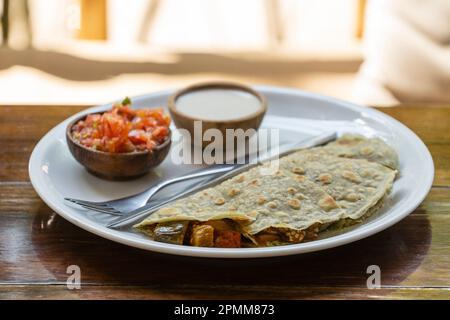 Quesadillas from spiruline tortilla with cashew cheese, eggplant, tomatoes and sour cream and tomato salsa on wooden table in tropical cafe, Thailand, Stock Photo