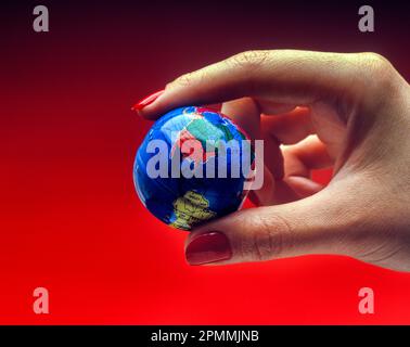 1991 HISTORICAL WOMAN’S HAND HOLDING TOY PLANET EARTH GLOBE ON RED BACKGROUND Stock Photo