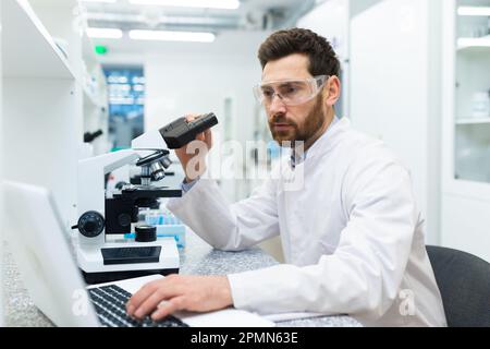 Serious and thoughtful scientist working in laboratory with microscope and laptop, mature experienced man in glasses and medical gown conducting experiments and entering data online into computer . Stock Photo