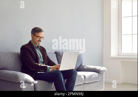 Happy successful mature entrepreneur working on laptop while sitting on sofa at home Stock Photo