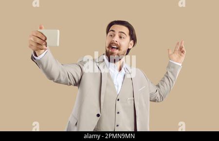 Cheerful stylish man in suit takes selfie, records video or talks online on beige background. Stock Photo