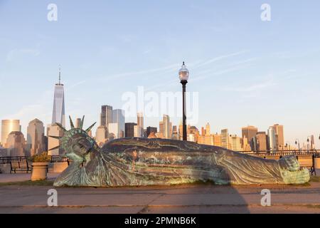 he sun sets on the 'Reclining Liberty' statue by artist Zaq Landsberg in Liberty State Park as it sits in front of the skyline of lower Manhattan and Stock Photo