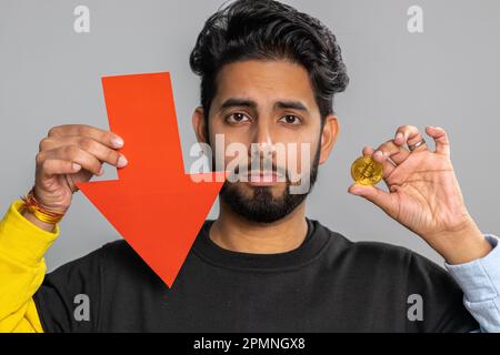 Upset indian man showing red arrow pointing down, downgrade, unsuccessful business, fall of cryptocurrency stock market investment, mining bitcoins BTC money exchange rate, bankruptcy fail. Hindu guy Stock Photo