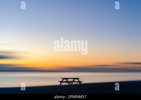 A lone bench seat on the beach at Bracklesham Bay looking over the Solent towards the Isle of Wight at sunset. Stock Photo