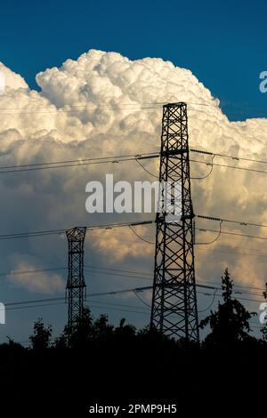 Silhouette of metal electrical towers with trees in the foreground and dramatic storm clouds in the background and blue sky above Stock Photo