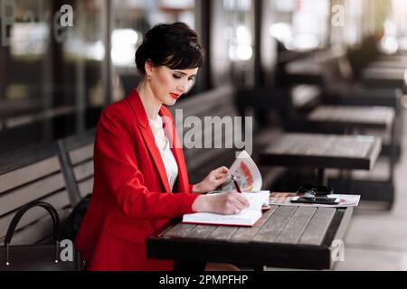 Serious pensive businesswoman making to do list and reading latest news in newspaper. Woman in red business blazer doing note in notepad at cafe terra Stock Photo