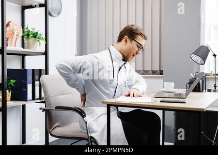 A young male doctor sits at his workplace and feels pain in his back. A doctor takes a break from his work day because of pain. Stock Photo