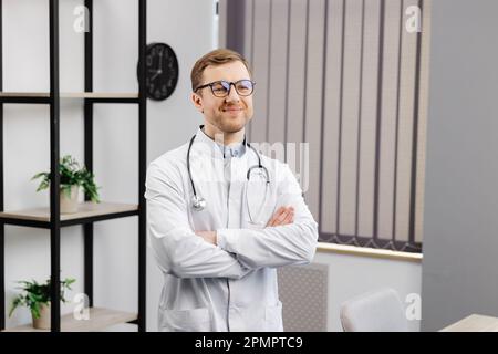 Smiling doctor posing with arms crossed in the office. Man is wearing a stethoscope and glasses. Medical staff on the workplace. Stock Photo