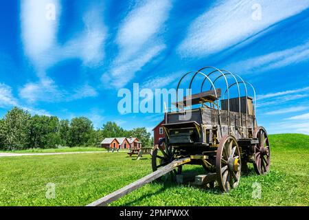 Old wooden wagon with dramatic clouds and blue sky with red painted buildings in the background, South of Longview, Alberta; Alberta, Canada Stock Photo
