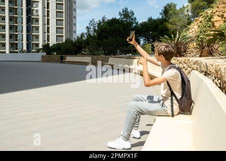 Happy teenager boy sitting on bench outdoors and taking selfie using smartphone app. Kid taking photo, making video call, communicate with his friends Stock Photo