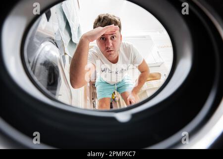 Man view from washing machine inside. Male does laundry daily routine. Hand over forehead looking far away, watching or searching. Stock Photo