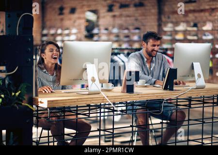 Young workers in a shop working on the computer and laughing, having a good time at work. Colleagues, teamwork, internet cafe concept. Stock Photo