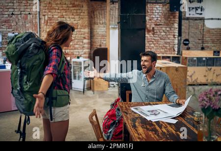 Handsome young man sitting in cafe meets friend, young woman with backpack standing with arms open, excited to see a friend. Meeting, lifestyle, toget Stock Photo