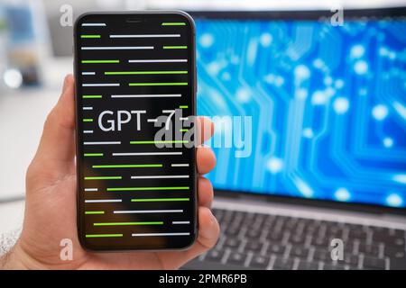 A mans hand holding a smartphone with GPT 7 neural network logo on the screen and a laptop displaying a neuronet on the splash screen in the background. March 2023, Prague, Czech Republic  Stock Photo