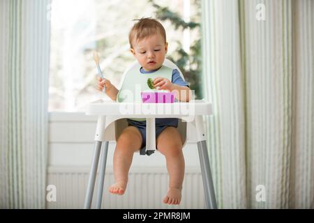 Cute baby eating first solid food, infant sitting in high chair. Child tasting vegetables at the table, discovering new food. Cozy kitchen interior. Stock Photo