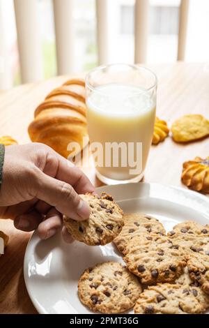 Taking a delicious sweet chocolate chip cookie from a plate, table setting with glass of milk and bread, pastry table, studio wallpaper Stock Photo