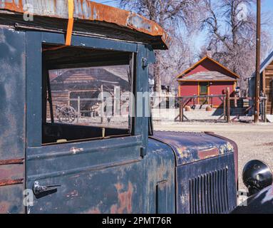 Looking through the window of a vintage antique truck you see old log cabins and homes on a dirt road. See door frame, radiator, head lamp closeup. Stock Photo