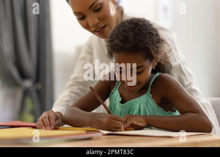 Smiling biracial mother sitting at dining table helping her daughter with school work Stock Photo