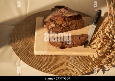 Fresh, brown, freshly baked, multi-grain and yeast-free bread on a cutting board and cereals, cut off a piece of the bread separately, the knife and copy space. Sun lights and shadows. High quality Stock Photo