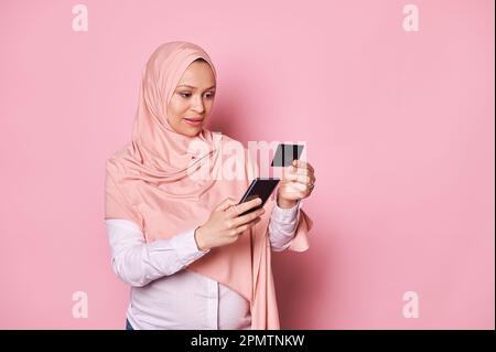 Happy pregnant Muslim woman in hijab, holding credit card and smartphone. Mobile apps. Internet banking. Online shopping Stock Photo