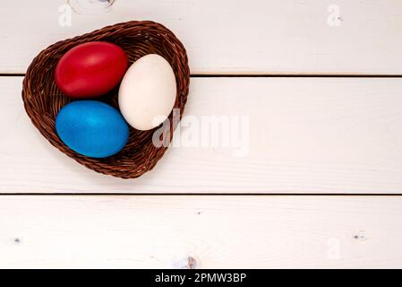 Multicolor decorated Easter eggs in the wicker bowl on a white wooden table. Easter concept. Stock Photo