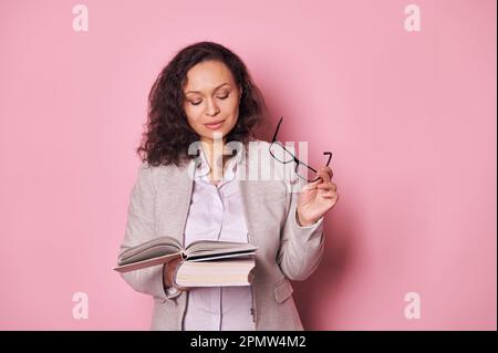 Curly haired brunette adult woman holding eyeglasses in her hand, reading studying book over pink background Stock Photo