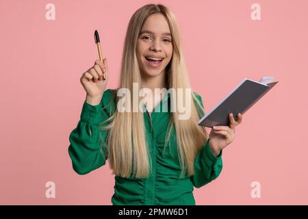 Thoughtful journalist caucasian woman making notes, writing down thoughts with pen into notepad notebook diary, to do list, good idea. Pretty teenager girl isolated alone on pink studio background Stock Photo