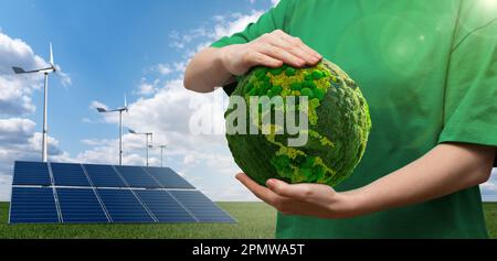 Woman holding a green planet Earth on a background of wind turbines and solar panels. Stock Photo