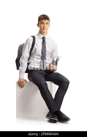 Male student in a school uniform holding books and sitting on a white cube isolated on white background Stock Photo