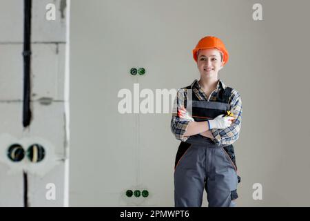 Portrait of a young professional woman electrician in a hard hat and overalls holding pliers for work, on a gray background. Stock Photo