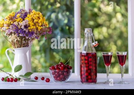 Homemade cherry brandy in two glasses and in a glass bottle on the windowsill by the window of the house on a summer day near the garden, close up Stock Photo