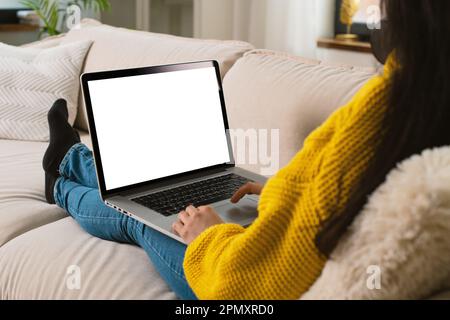 Unrecognizable multiracial woman in a yellow sweater in a modern living room lying on sofa working with a blank screen modern laptop computer mock-up Stock Photo