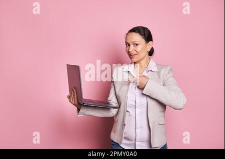 Charming African American business woman manager points at laptop computer, smiles looking at camera, isolated on pink Stock Photo