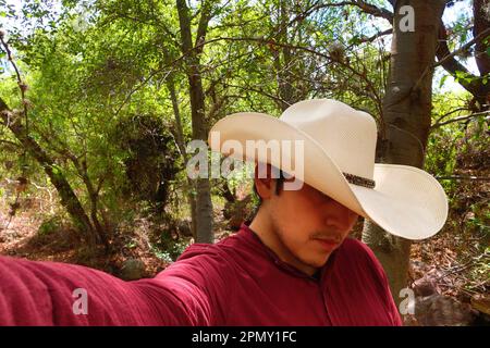 A man in a red shirt and a white cowboy hat is taking a selfie. Stock Photo