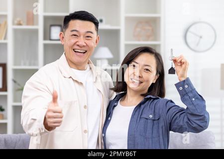 Happy young Asian couple bought an apartment, a house. They stand hugging and smiling at the camera. The man shows super with his finger, the woman holds the keys in her hand. Stock Photo