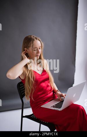 A happy business woman blogger in a red dress is working, typing on a laptop apple and sitting on a chair. portrait blonde assistant of hands with computer MacBook . Vertical Stock Photo
